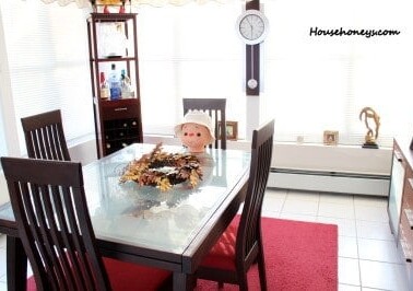 A dining room table with a doll sitting in a chair.