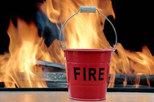 A red fire bucket in front of a burning fire.