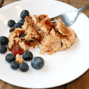 A baked scoop of Dump Cake on a white plate with blueberries around it.