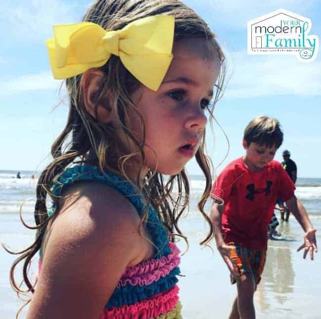 A little girl at the beach with a little boy walking past her.