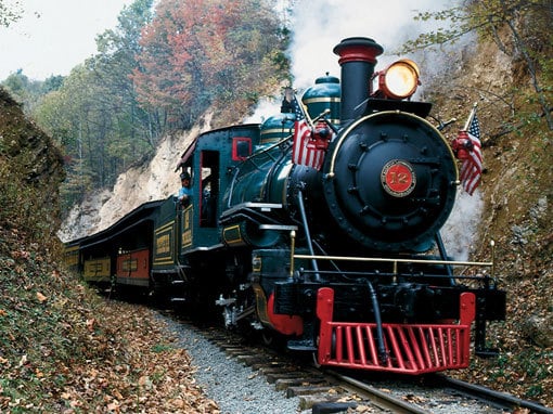 A steam engine train traveling down train tracks near a forest.