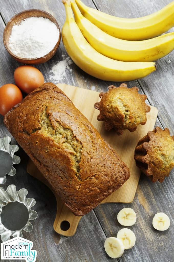 A loaf of banana bread with banana cupcakes beside it with bananas resting on the table.