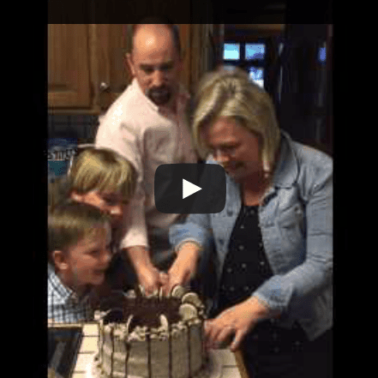 A video clip of a family watching a woman cutting a cake.