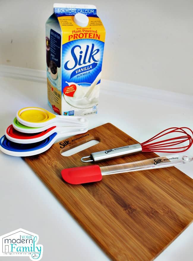 A cutting board with a spatula, whisk and measuring spoons and a container of Silk Milk.