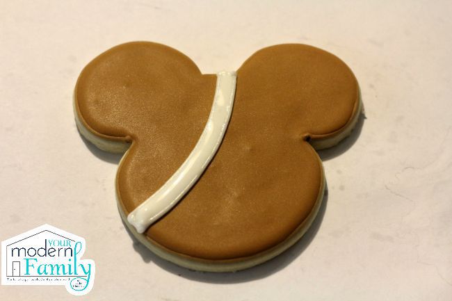 A Mickey Mouse shaped cookie with brown icing and a white stripe on it.