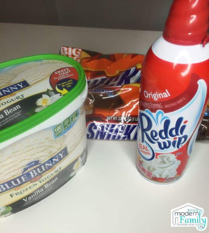 A container of ice cream with a bag of Snickers and a container of Reddi-wip beside them.
