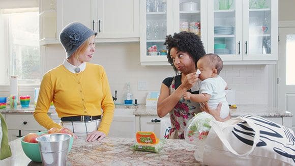 Two women in a kitchen with one woman holding a baby while wiping his nose with Boogie Wipes.