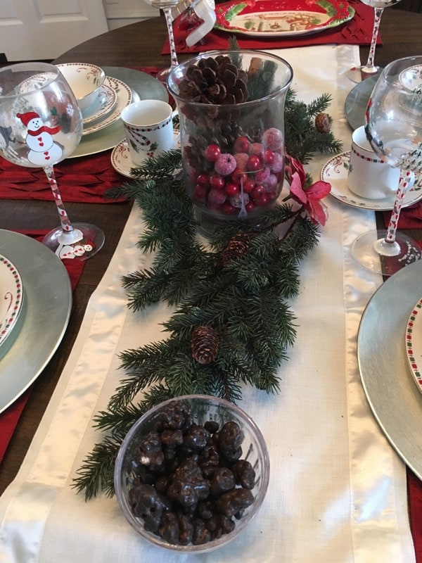 A dining room table set with Christmas dishes and glasses with pine cones in a glass vase.