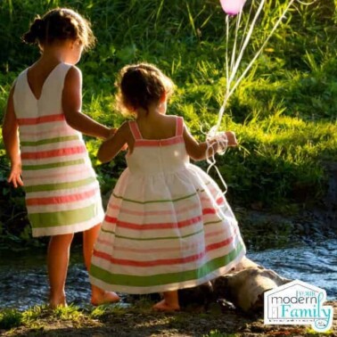 Two little girls in dresses standing near a creek while they hold balloons.