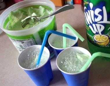 Three blue cups with soda and lime sherbet and a straw with a bottle of soda and a tub of sherbet behind them.