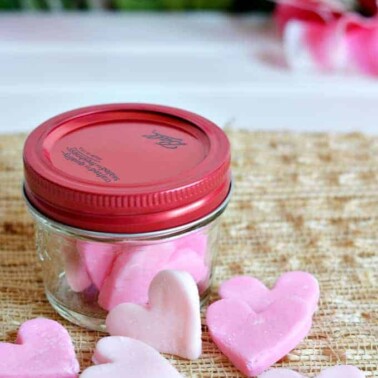 A small glass jar with a red lid and candy hearts around it.