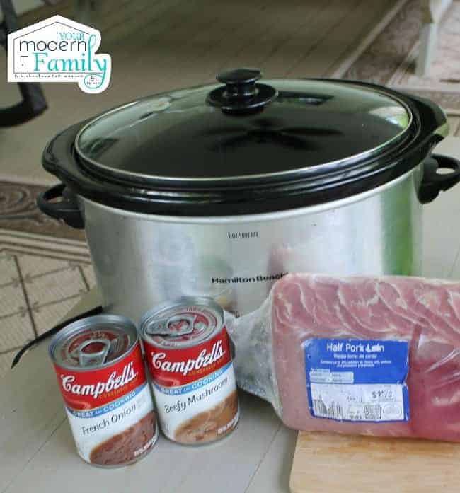 A silver crock pot with two cans of Campbell soup and a pork loin resting in front of it.