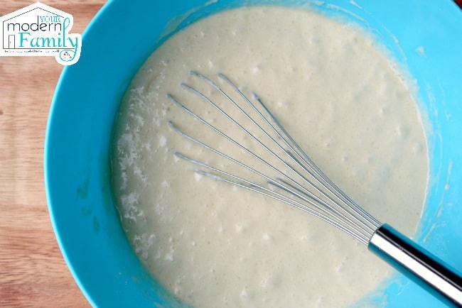 A blue bowl on a table with white batter and a whisk in it.