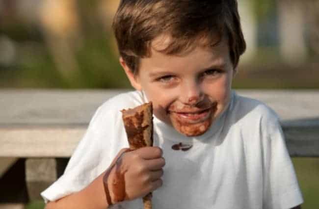 A boy eating an ice cream cone with it melting on his face, hand and shirt.