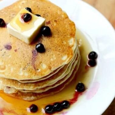 A stack of Blueberry pancakes with syrup and butter on a white plate.