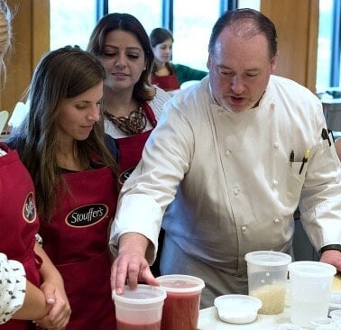 A group of people standing around a table with a chef showing ingredients.