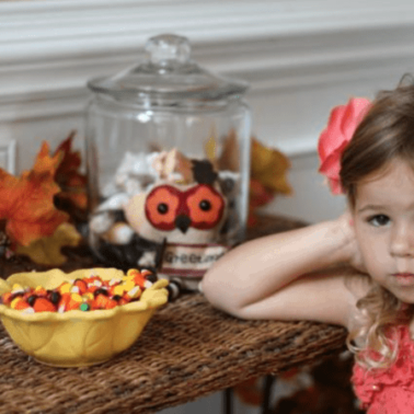 A little girl sitting at a table with a bowl of Halloween candy and a glass jar behind her.