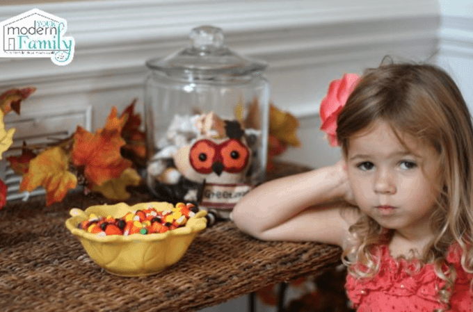 A little girl sitting at a table with a bowl of Halloween candy and a glass jar behind her.