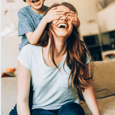 A boy standing behind a woman sitting on the floor with his hands over her eyes.