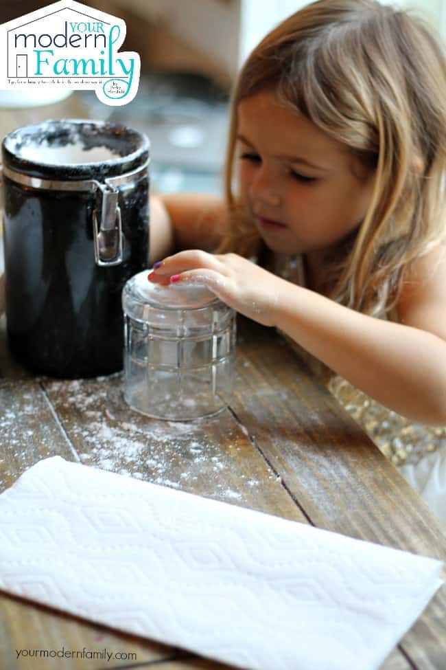 A little girl sitting at a table with a container of flour in front of her.