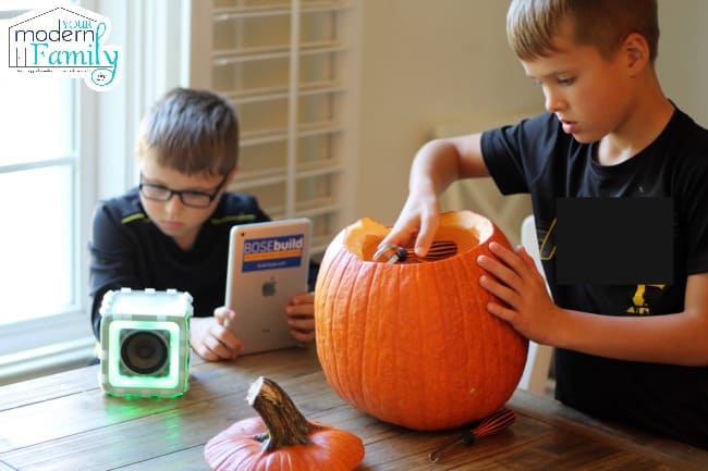 Two boys sitting on a table cutting out a pumpkin with a Bose build speaker cube.