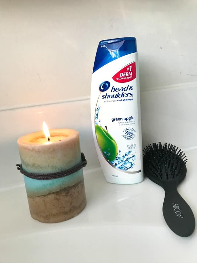 A close up of a bottle of Head and Shoulders with a brush and a candle beside it.
