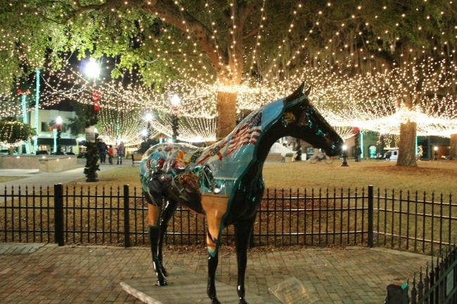 A statue of a horse that is standing in front of a fence with Christmas lights in the background.