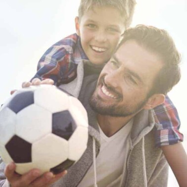 A man holding a boy on his shoulders and a soccer ball.