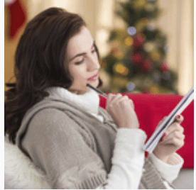 A woman sitting on a couch holding a pen and note pad with a Christmas tree in the background.