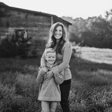 A woman and a little boy standing in a grassy field with a barn behind them.