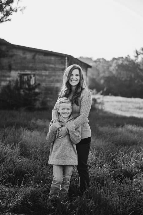 A woman and a little boy standing in a grassy field with a barn behind them.
