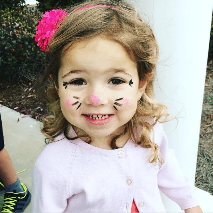 A little girl posing for a picture with her face painted to look like a cat.