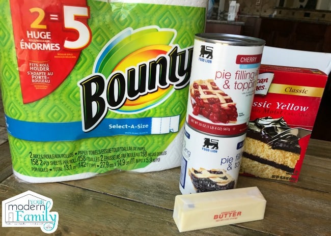 A cake mix box with two cans of pie filling, a stick of butter and a package of Bounty paper towels on a table.