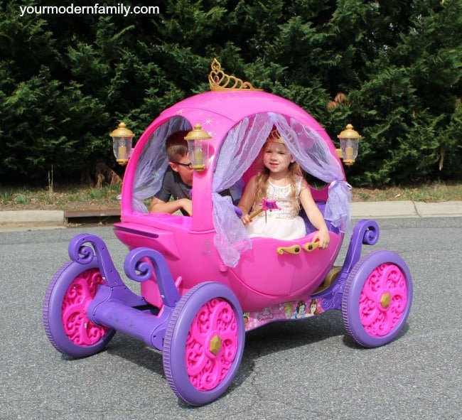 A little girl riding in a pink carriage child\'s ride on toy with a boy driving it.