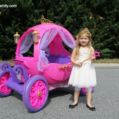 A little girl dressed as a princess standing by a child's carriage theme ride on toy.