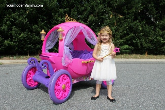 A little girl dressed as a princess standing by a child\'s carriage theme ride on toy.