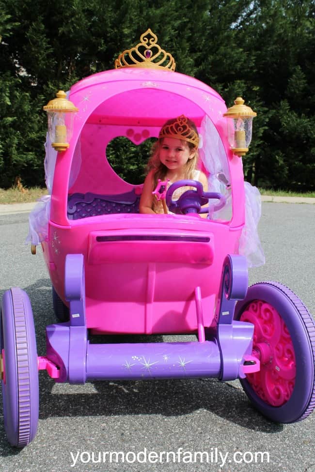A close up of little girl with a crown on her head driving a pink carriage ride on toy.