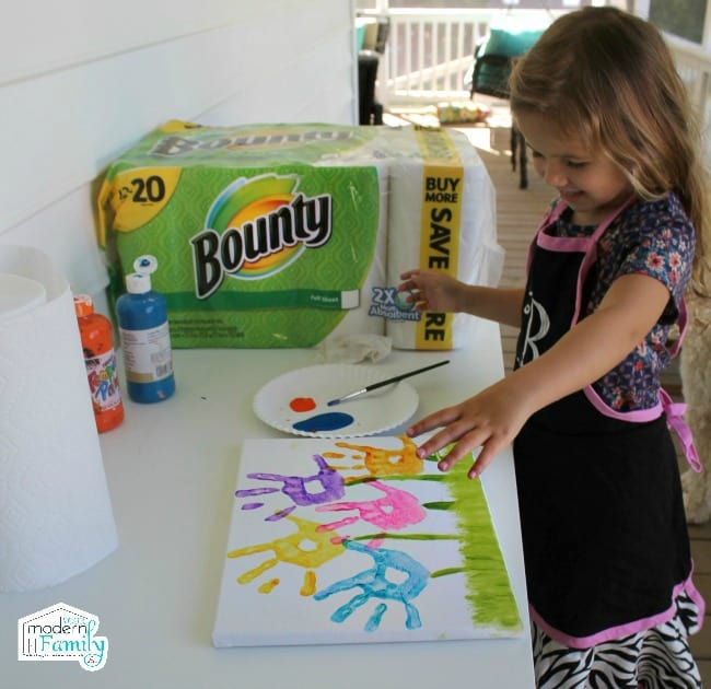 A little girl standing at a counter painting a picture with a package of Bounty paper towels beside her.
