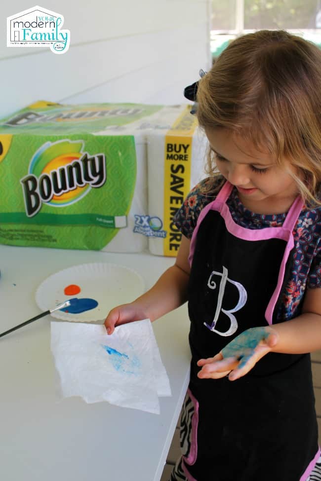 A little girl standing at a counter with paint on her hand with a package of Bounty paper towels beside her.