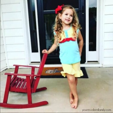 A girl posing in front of a window leaning on a rocking chair.