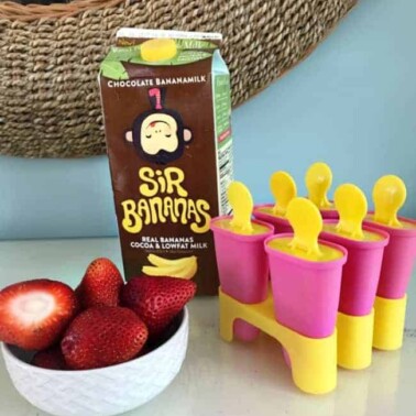 A bowl of strawberries and a container of chocolate milk beside freezer pop molds.