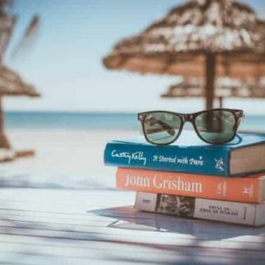 A stack of books with sunglasses resting on them with beach umbrellas and ocean behind them.