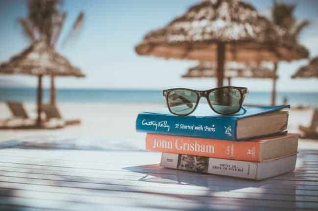 A stack of books with sunglasses resting on them with beach umbrellas and ocean behind them.