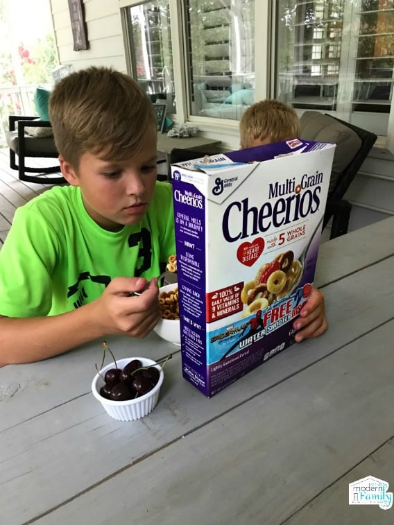 A young boy sitting at a table eating while he reads the back of a cereal box.
