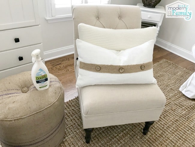 A spray bottle of Downy sitting on a ottoman beside a cushioned chair with a pillow on it.