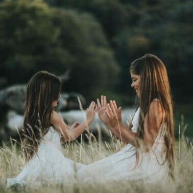 Two girls in white dresses kneeling in a field touching hands.