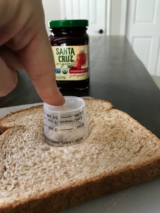 A finger pressing a plastic lid into a slice of bread with a jar of jelly behind it.