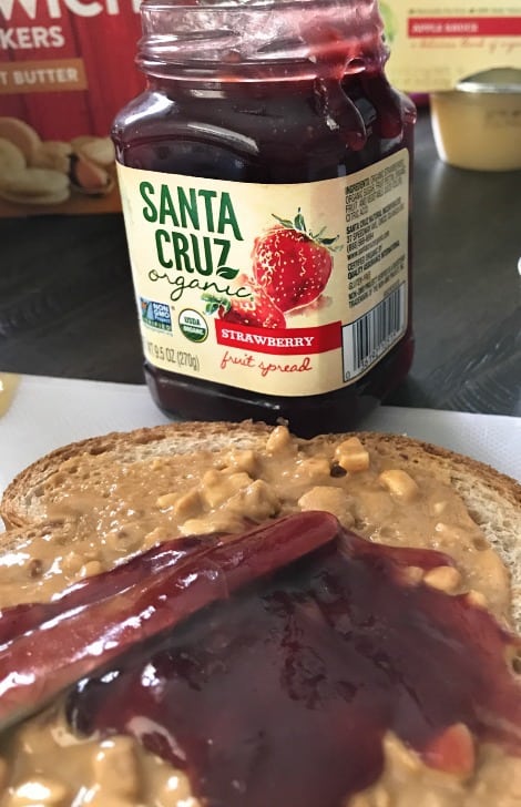 A close up of a knife spreading jelly over a slice of peanut butter bread with a jar of jelly.