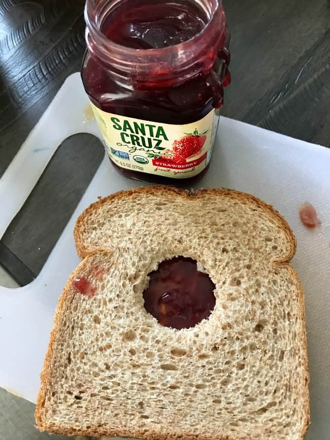 A sandwich with a hole cut in the top slice of bread showing jelly in the hole.