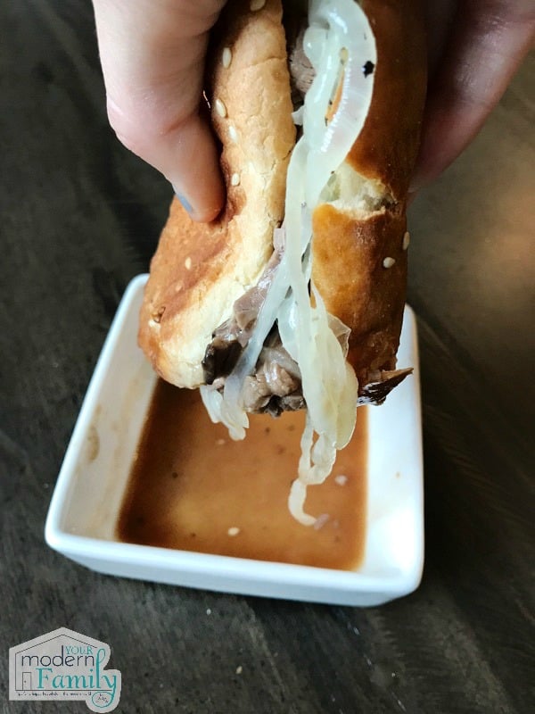 A hot roast beef sandwich in someones hand with a bowl of dipping gravy on the table.